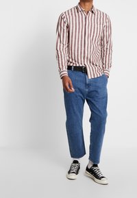 Langsleeve shirt met rode en witte strepen, gecombineerd met blauwe denim jeans. Een zwarte riem en zwarte sneakers met witte accenten maken de look compleet.