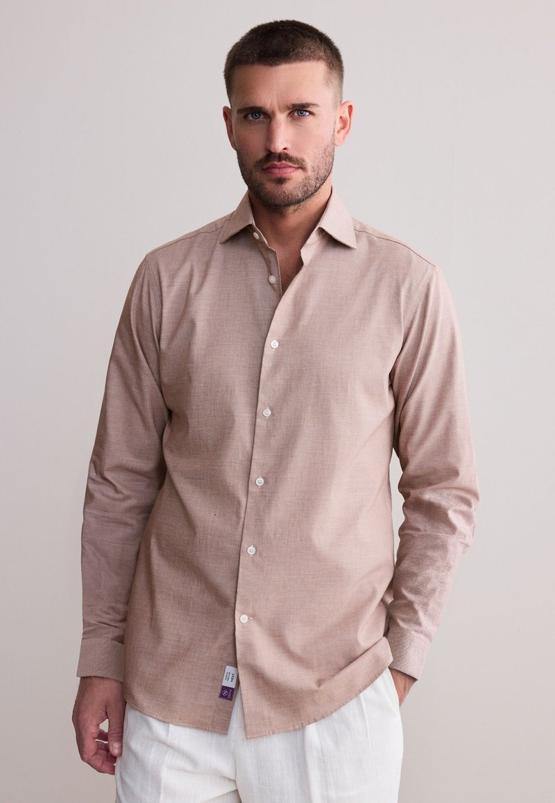 Light brown long-sleeve button-up shirt with a subtle texture, white buttons, and a curved hem, paired with white trousers.