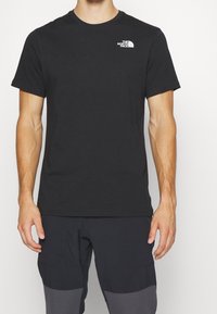 Hombre que lleva una camiseta negra de manga corta sencilla con el logo de The North Face en el pecho, combinada con pantalones negros y grises.