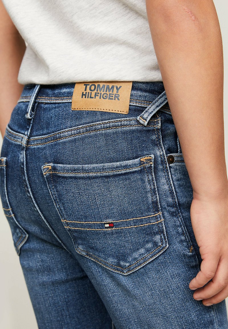 Dżinsy w średnim niebieskim kolorze z kontrastującym pomarańczowym szwem. Posiadają skórzaną metkę z napisem "TOMMY HILFIGER" oraz drobny detal logo na kieszeni.