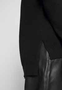 Schwarzer Pullover mit strukturiertem Finish und ribbed Saum, teilweise in schwarze Kunstlederhose gesteckt, mit einem diagonalen Seitenschlitz.