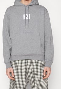 Sweat à capuche gris avec cordon de serrage, arborant un petit logo jumpman noir sur un patch blanc. Assorti à un pantalon à carreaux verts et blancs.