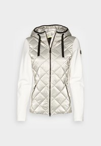 Prošívaná stříbrná vesta s kapucí, černými šňůrkami a bílými dlouhými rukávy. Má přední zip a boční kapsy.