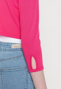 Hot pink cropped shirt with three-quarter sleeves, featuring a decorative cutout detail at the cuffs, paired with light blue denim.
