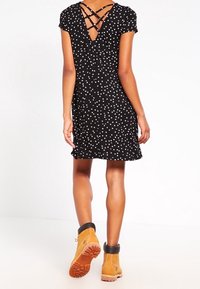 Musta polkadot-kuvioinen mekko, jossa on V-muotoinen selkä ja ristikkäiset olkaimet. Yhdistetty ruskeisiin nilkkureihin, joissa on musta resori ja oranssi pohja.