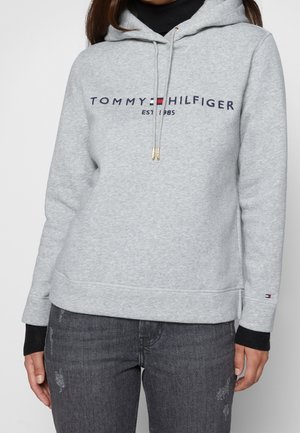 Osoba ubrana w jasnoszarą bluzę z kapturem Tommy Hilfiger z logo i napisem "EST 1985", zestawioną z ciemnoszarymi podartymi dżinsami oraz czarnymi wewnętrznymi rękawami.