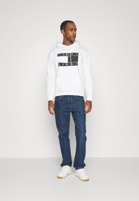 Sudadera blanca con el logo negro de "TOMMY JEANS", jeans azules y zapatillas blancas con suelas de goma. El modelo está de pie sobre un fondo sencillo.