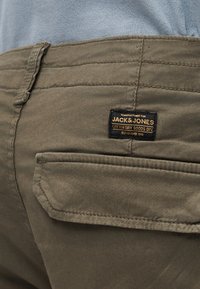 Κοντινό σε τσέπη χακί παντελονιού με μαύρη και χρυσή ετικέτα της μάρκας Jack & Jones και θηλιά ζώνης από πάνω.
