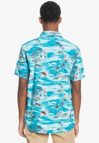 Lyhythihainen paita turkoosissa kankaassa, jossa on trooppinen kuosi palmuilla, purjelaivoilla ja pilvillä. Napillinen kaulus ja väljä istuvuus.