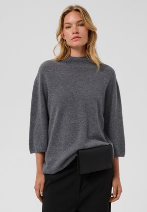 Donna con capelli biondi che indossa un maglione grigio largo e pantaloni neri con una pochette nera rettangolare alla vita.