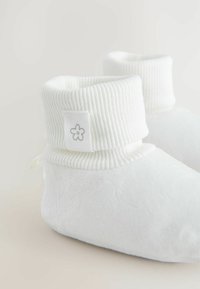 Bottillons en polaire blanche avec un revers côtelé, arborant une petite étiquette en tissu avec un motif de fleur. Texture douce et forme arrondie.