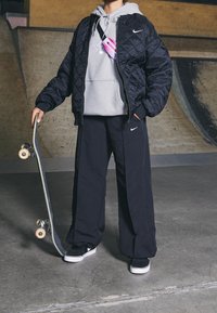 Persona de pie en un skatepark sosteniendo una patineta, vistiendo una chaqueta acolchada negra, sudadera gris, pantalones anchos negros y zapatillas negras con suelas blancas.