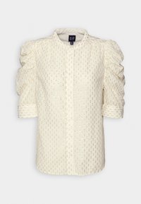 Camicia cremosa con bottoni e maniche a sbuffo, caratterizzata da un motivo testurizzato di piccole macchie dorate. Tessuto morbido con una leggera lucentezza.