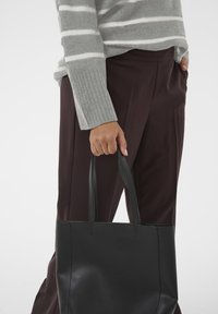 Czarna skórzana torba noszona w ręce, o gładkiej fakturze i minimalistycznym wzornictwie. Szary sweter w paski i ciemne spodnie częściowo widoczne.