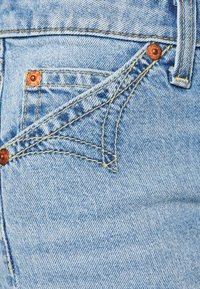 Vaaleansiniset farkut, joissa on pieni etutasku, kuparin niittejä ja erottuvaa keltaista ommelta, esittelevät teksturoitua kangasta.