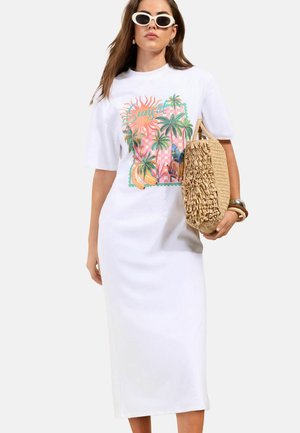 Žena v bielych midi šatách s farebnou potlačou západu slnka a palmy, biele slnečné okuliare, drží textúrovanú tkanú slamennú kabelku.