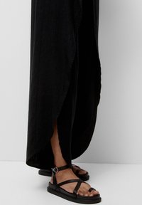 Robe maxi noire fluide avec une fente latérale, associée à des sandales plates noires à bretelles croisées et bride de cheville avec boucle.