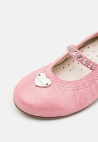 Chaussure d’enfant Mary Jane rose pailletée avec une bride ornée de bijoux et un pendentif en forme de cœur argenté sur l’orteil.