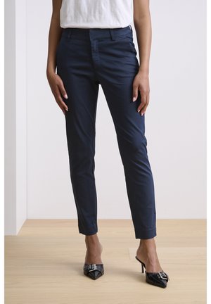 Frau trägt schlanke marineblaue Hose und schwarze spitze High Heels mit Schnallendetail, steht auf hellem Holzboden.