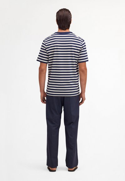Uomo in piedi di spalle che indossa una maglietta a righe navy e bianche a maniche corte, pantaloni navy e sandali neri su sfondo bianco.