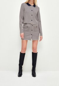 Kvinna iförd en rutmönstrad långärmad knappljacket och matchande mini-kjol med framfickor, kombinerat med svarta knähöga stövlar.