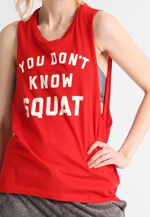 Person iført en rød ærmeløs skjorte med hvid tekst "YOU DON'T KNOW SQUAT" og grå bukser, hånd hvilende på hoften.