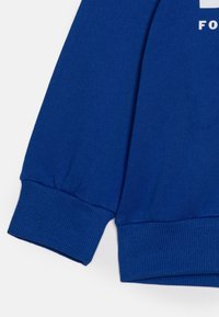 Modrá mikina s vroubkovanými manžetami a hladkým povrchem. Design zahrnuje bílé písmo na přední straně. Materiál je měkký a pružný.