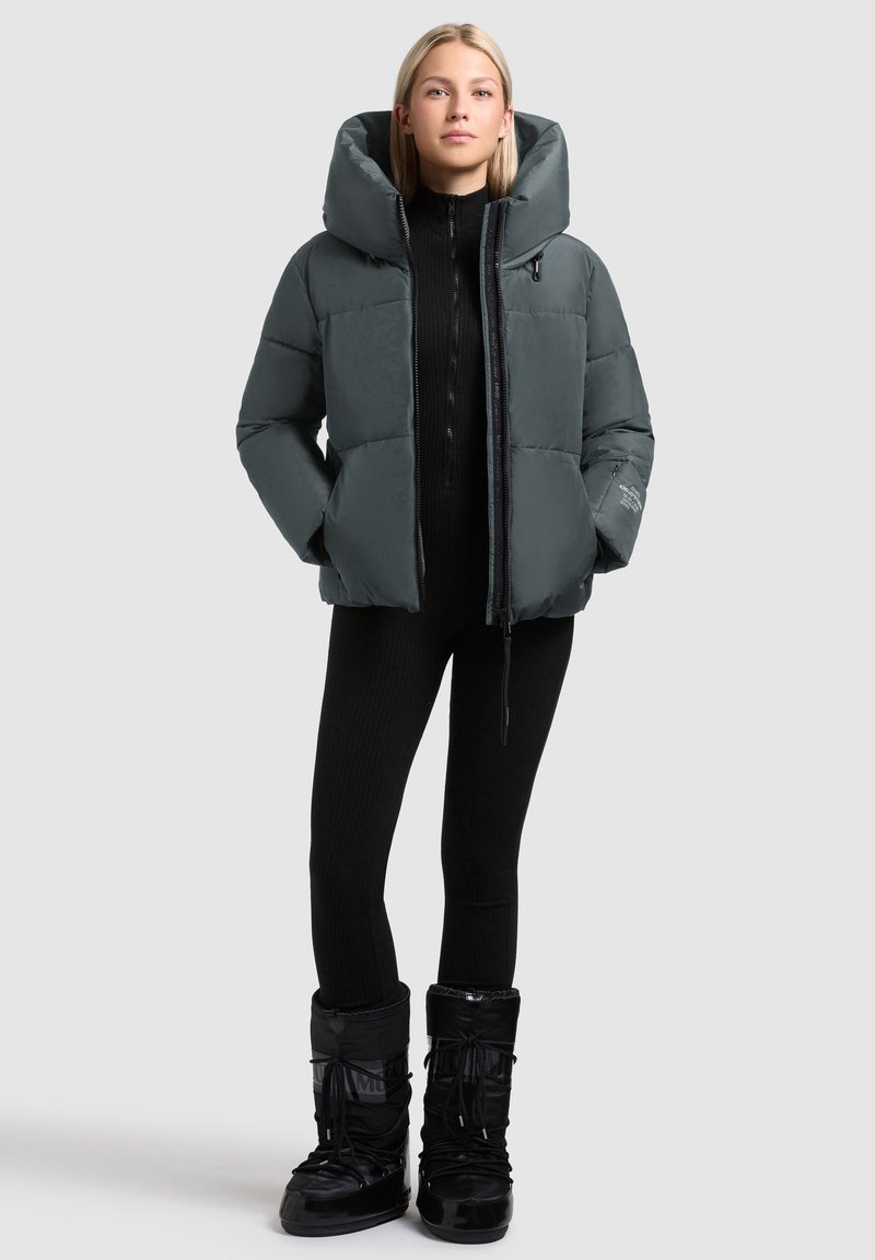 Ung kvinde iført en grå polstret jakke, sorte ribbede leggings og sorte vinterstøvler, stående med hænderne i lommerne mod en lys baggrund.