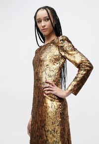 Vestido de mangas compridas cintilante feito de lantejoulas douradas e bronze, com silhueta ajustada e detalhes texturizados na superfície.