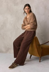 Brauner Strickcardigan mit V-Ausschnitt, kombiniert mit weiten braunen Hose. Das Model trägt braune Sneaker. Im Hintergrund ist ein gelber Akzentstuhl zu sehen.