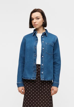 Ung kvinne med skulderlangt mørkt hår iført en åpen blå denimskjorte over en hvit topp og et svart skjørt med hvite polkadotter.