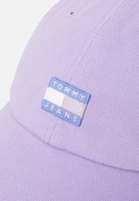 Lilla bomullslue med strukturert overflate, med en rektangulær blå lapp som viser "TOMMY JEANS" i hvit og lyseblå skrift.