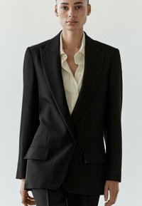 Czarna, szyta na miarę marynarka z szpiczastymi klapami, dwie przednie kieszenie, gładka faktura, w połączeniu z jasnobeżową koszulą na guziki pod spodem.