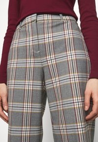 Grå rutiga byxor med mönster i vitt, gult och burgunder, med rak passform och en slät tygtextur. Accentdetalj på fickan synlig.