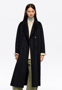 Mujer con abrigo negro de doble botonadura, camisa beige, suéter de cuello alto gris y pantalones oliva, de pie contra un fondo blanco.