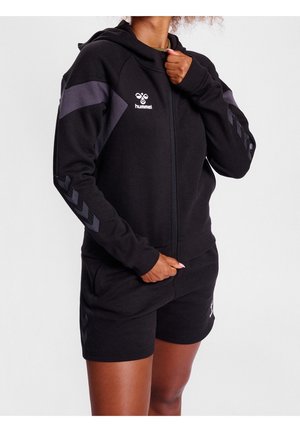 Person iført sort hummel lynlås-hættetrøje med chevron-mønstre på ærmerne og matchende sorte shorts mod hvid baggrund.