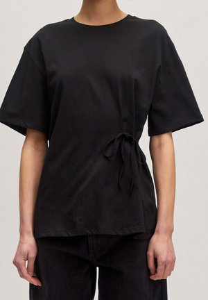 Personne portant un t-shirt noir à manches courtes avec un détail noué sur le côté, associé à un pantalon noir, debout devant un fond uni.