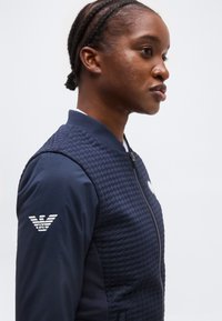 Προφίλ ατόμου με πλεξούδες που φοράει μια μπλε σκούρη καπιτονέ ζακέτα με λευκό λογότυπο στο μανίκι, σε απλό φόντο.