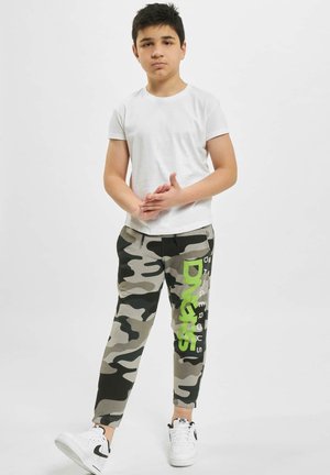 Grau getarnte Jogginghosen mit grünem Text, kombiniert mit einem schlichten weißen T-Shirt und weißen Sneakers mit schwarzen Akzenten.