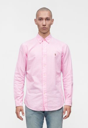 Homme de face portant une chemise rose à rayures verticales et un jean bleu clair sur un fond blanc uni.