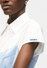 Κοντινό πλάνο ενός ατόμου που φοράει άσπρο κοντομάνικο πουκάμισο με μπλε σχέδιο και τη λέξη «MAX&Co.» κεντημένη στο μανίκι.