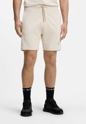 Mann trägt beige Shorts mit Kordelzug, schwarze Socken mit dem Schriftzug „BOSS“ und schwarze Sportschuhe, steht vor weißem Hintergrund.