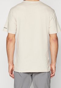 Béžové tričko s krátkým rukávem, vyrobené z měkké bavlny, s kulatým výstřihem a malým černým nápisem "Columbia" na levém rukávu.