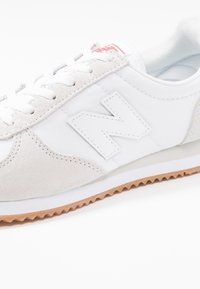 Chaussure de sport blanche en matériaux suède et tissu, avec un logo N blanc bien visible, des accents gris clair texturés et une semelle extérieure en caoutchouc couleur gomme.
