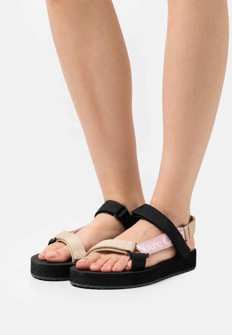Pieds portant des sandales à brides ajustables beige, noir et rose avec des semelles épaisses noires, posés sur une surface blanche.