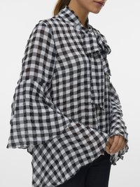 Γυναίκα που φοράει πουκάμισο ασπρόμαυρο με καρό σχέδιο, με μακριά μανίκια καμπάνα και δέσιμο φιόγκο στον λαιμό, στέκεται με τα χέρια ενωμένα.