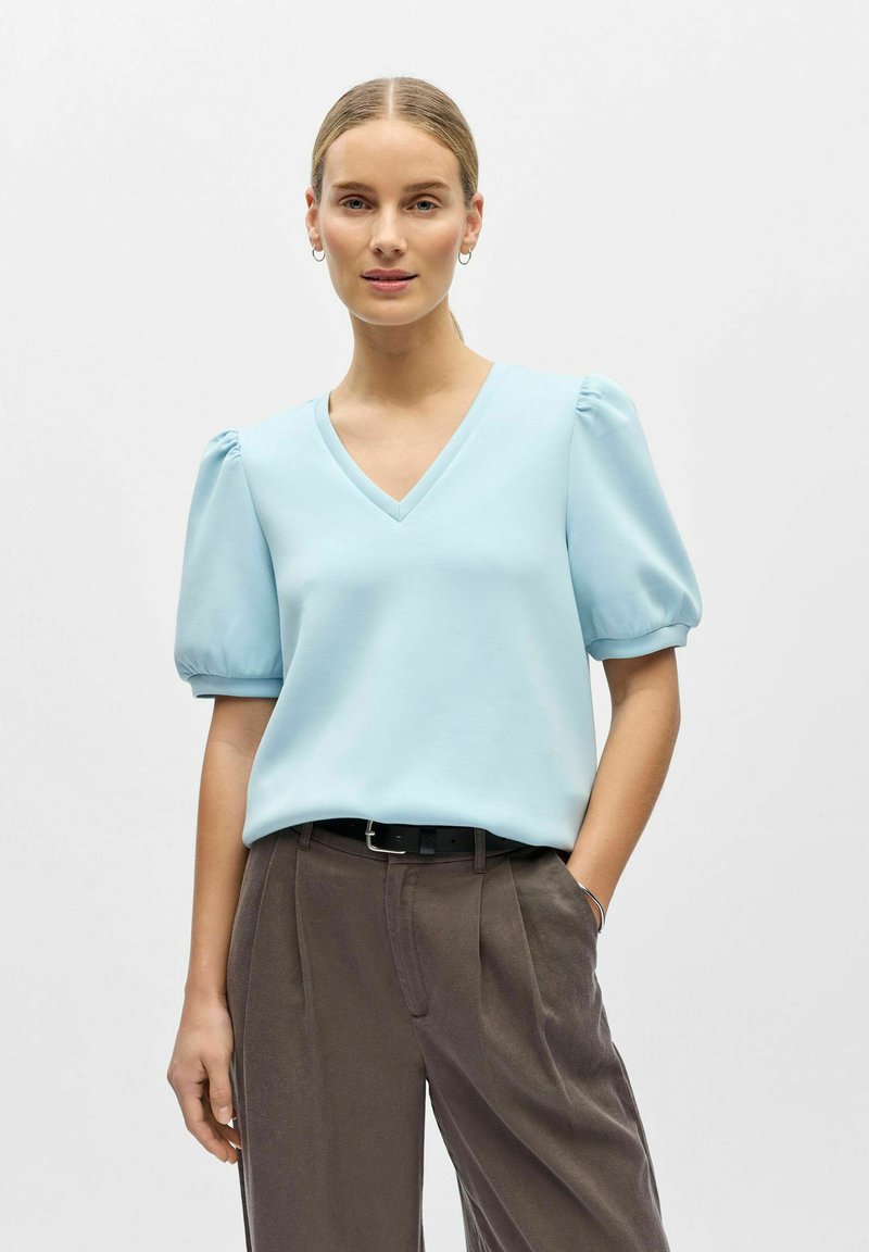 Haut bleu clair à manches courtes avec un col en V et des manches bouffantes, associé à un pantalon large marron foncé et une ceinture noire. Texture de tissu lisse.