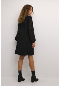 Black dress with sheer floral lace sleeves, slightly A-line cut, knee-length, paired with black ankle boots. Smooth texture with subtle shine.