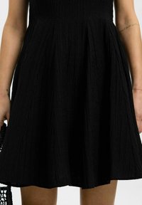 Musta mekko, jossa on teksturoitu, poimuuntunut kangas, istuva bodice ja leveä hame; yhdistettynä kudottuun mustaan laukkuun.