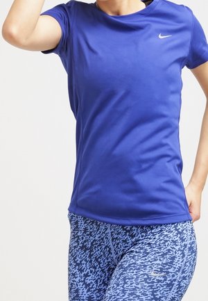 Henkilö pukeutuneena siniseen Nike-urheilupaitaan ja kuvioituihin sinisiin legginsseihin, poseeraa yhdellä kädellä koukussa ja kädellä pään lähellä.